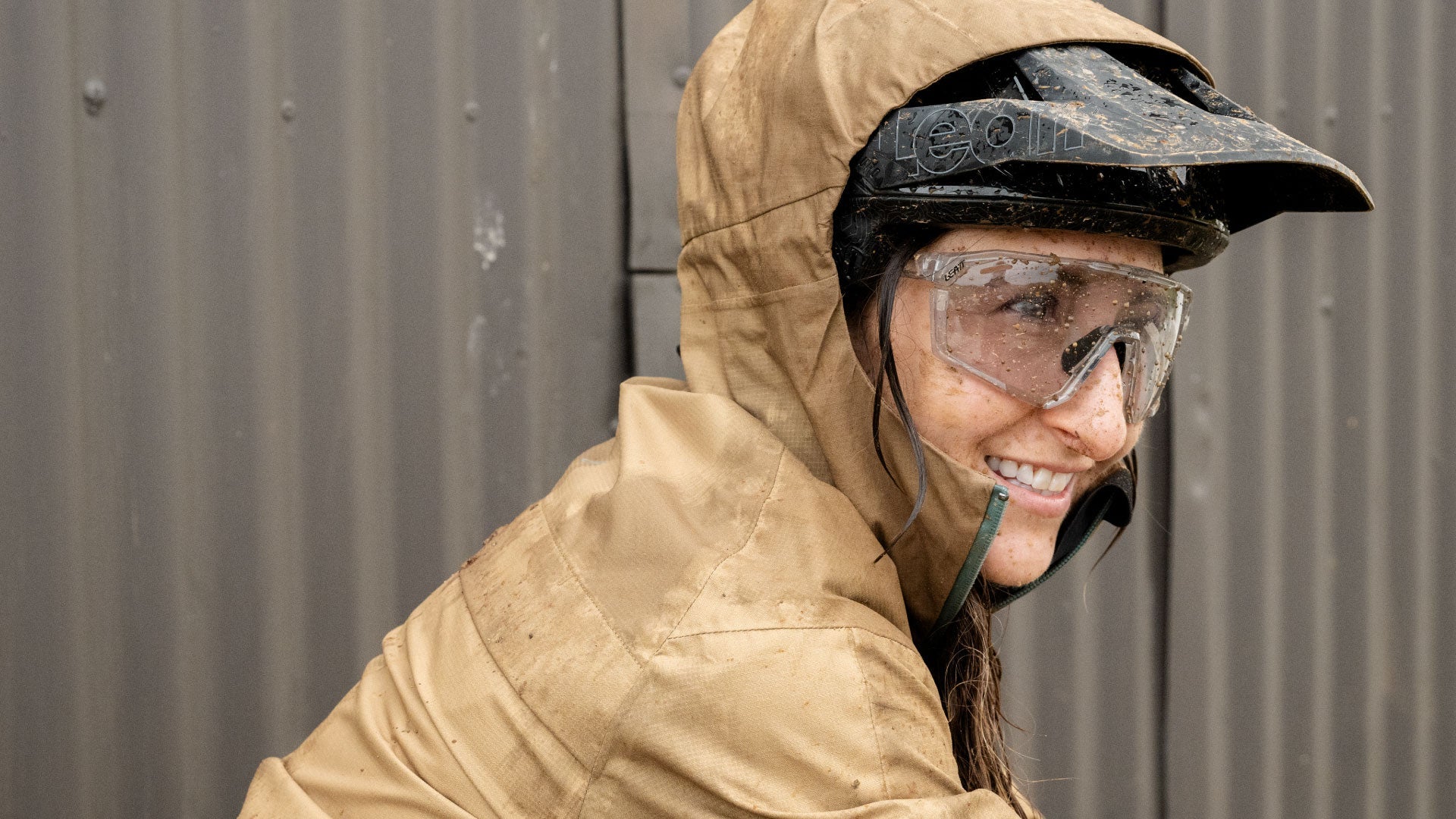 Person wearing a brown hooded jacket and black helmet with goggles, standing against a metallic wall.
