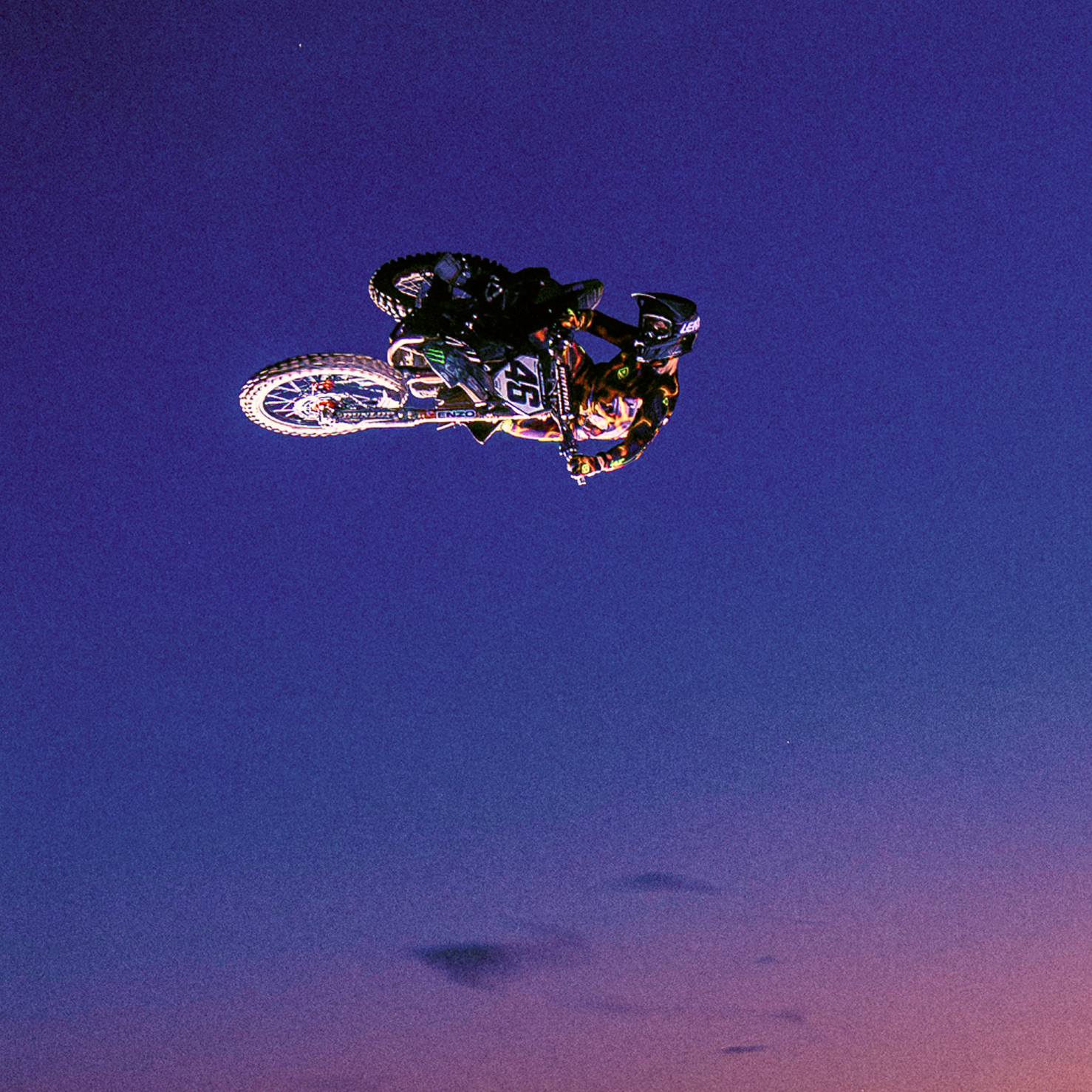 A dirt bike is captured mid-air performing a jump over a mountainous landscape at sunset, with a dramatic purple, orange, and blue sky background.