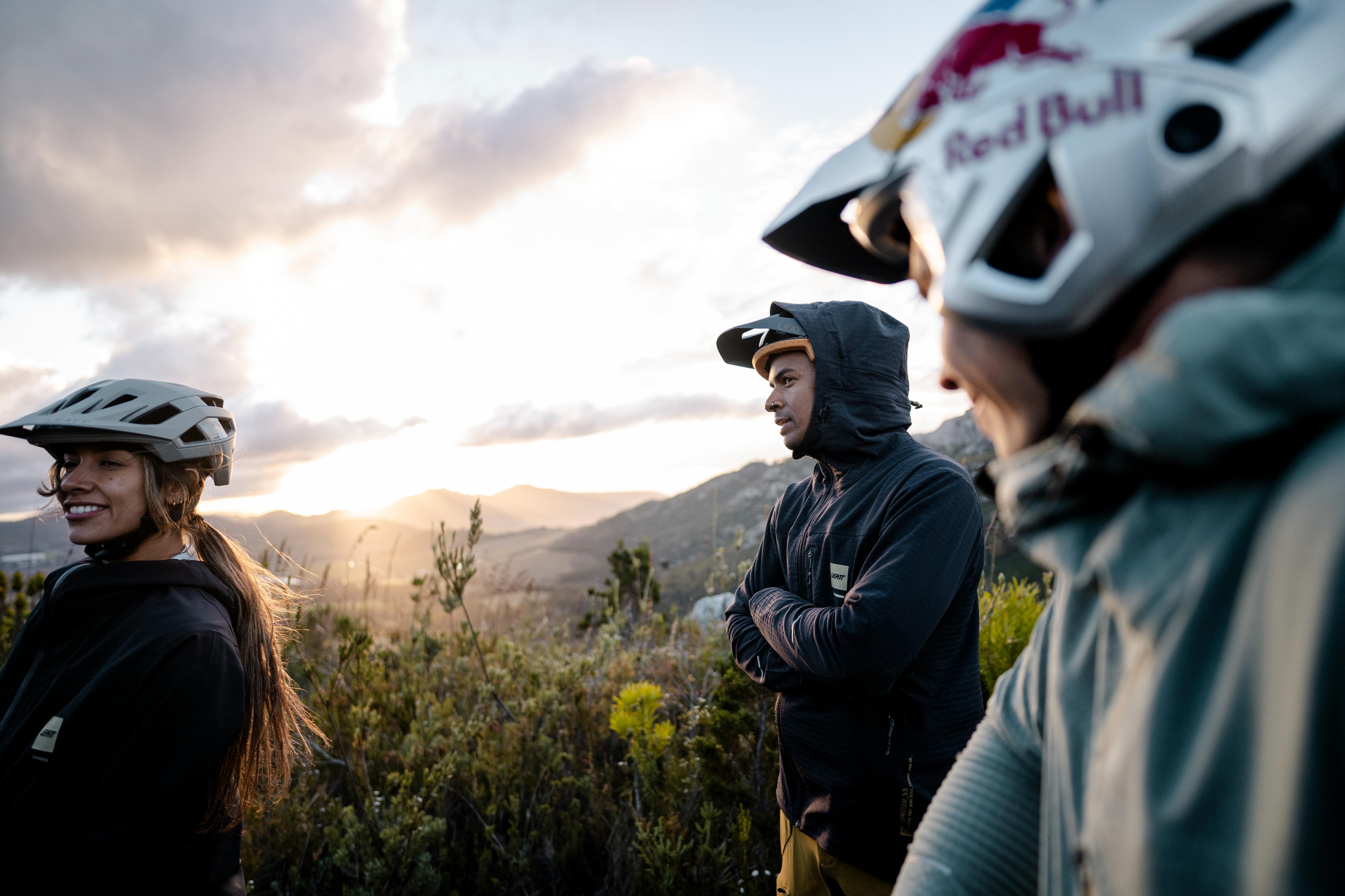Two people wearing helmets with a Red Bull logo, standing in a mountainous landscape during sunset.