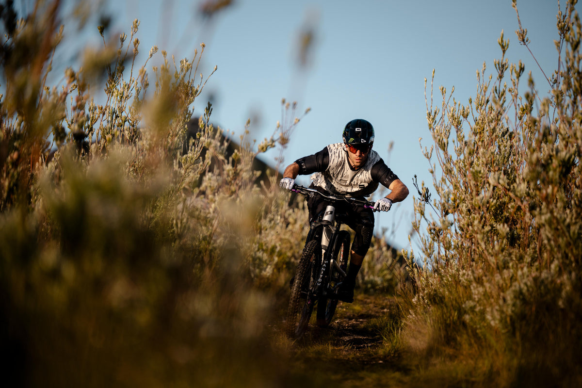 Person riding a mountain bike on a trail with tall grass and shrubs.