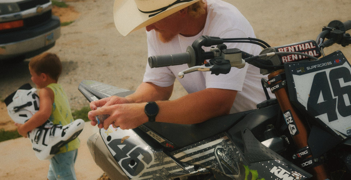 A man in a white cowboy hat and white t-shirt leans over a black and white number 46 dirt bike at a dirt track, while a young boy in a yellow shirt walks past carrying a white and black motocross boot. V