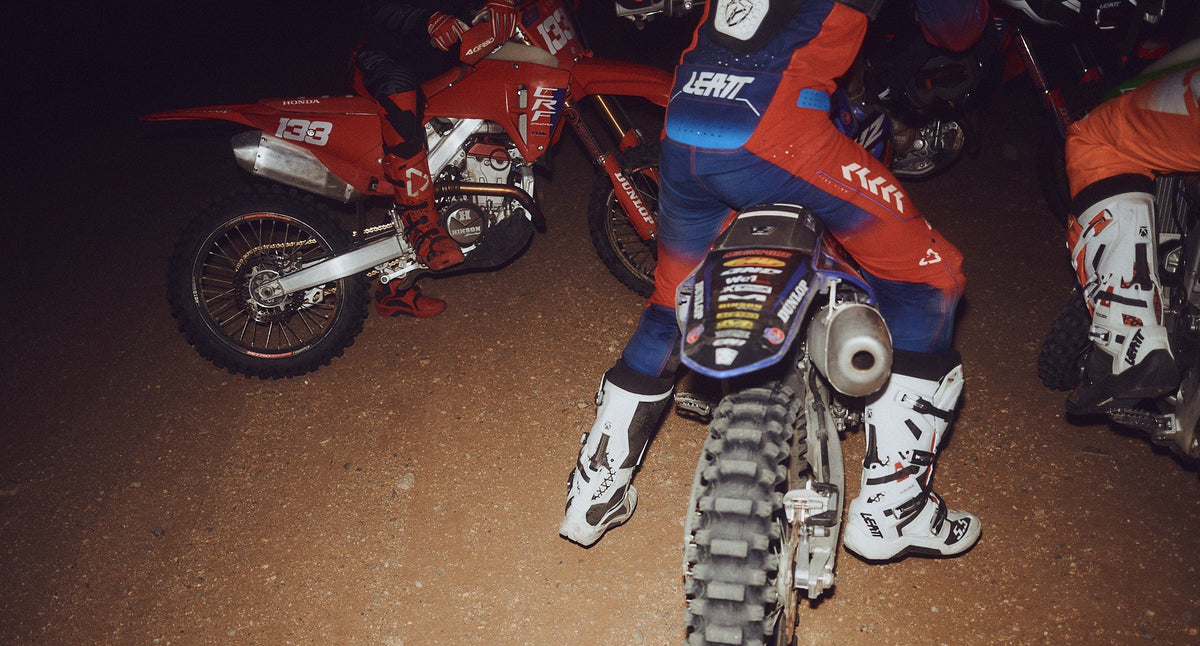 A motocross rider in blue, red, and white gear sits on a blue and black dirt bike at night on a dirt track, with other red and black bikes visible in the background.