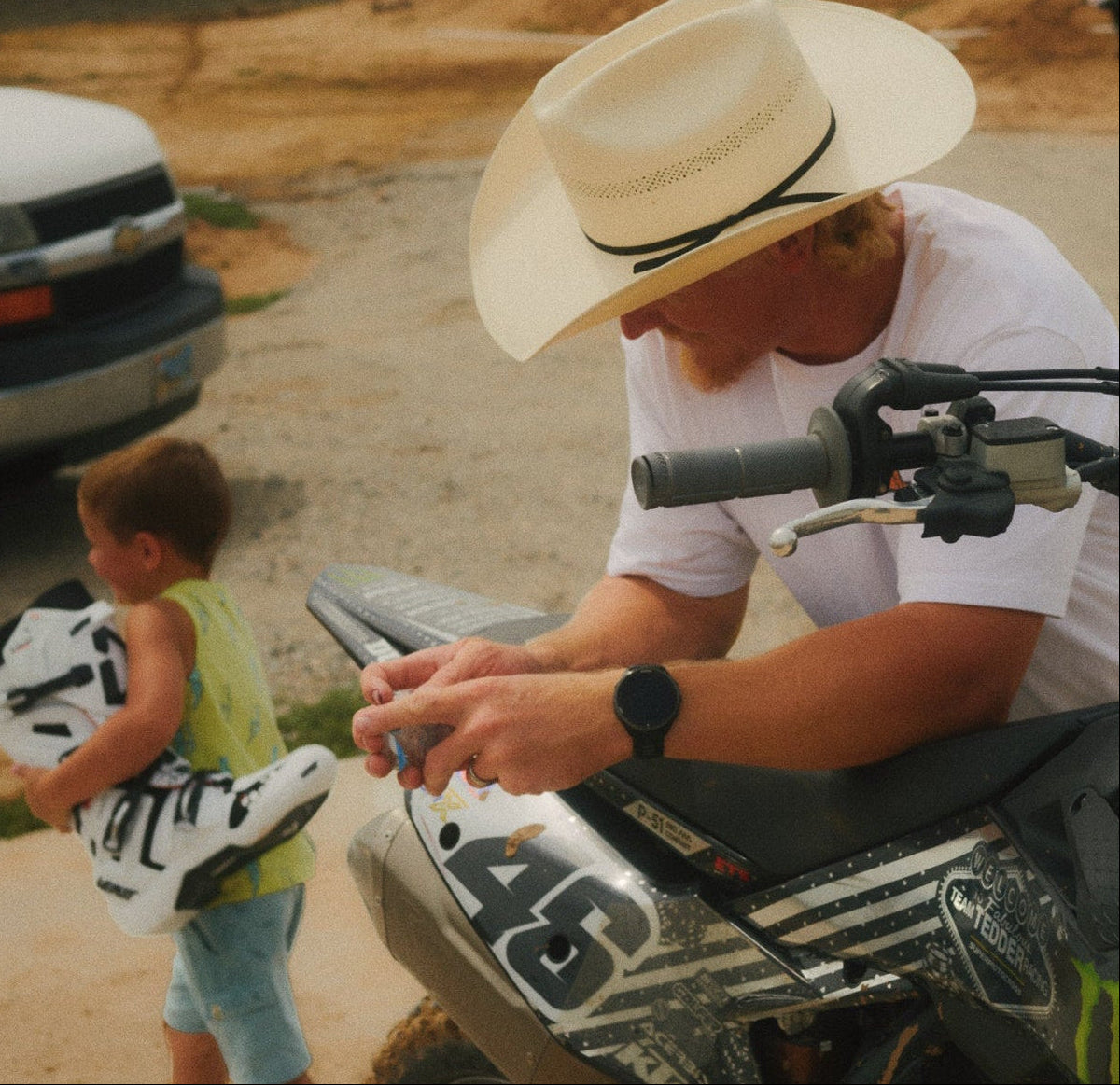 A man in a white cowboy hat and white t-shirt leans over a black and white number 46 dirt bike at a dirt track, while a young boy in a yellow shirt walks past carrying a white and black motocross boot. V