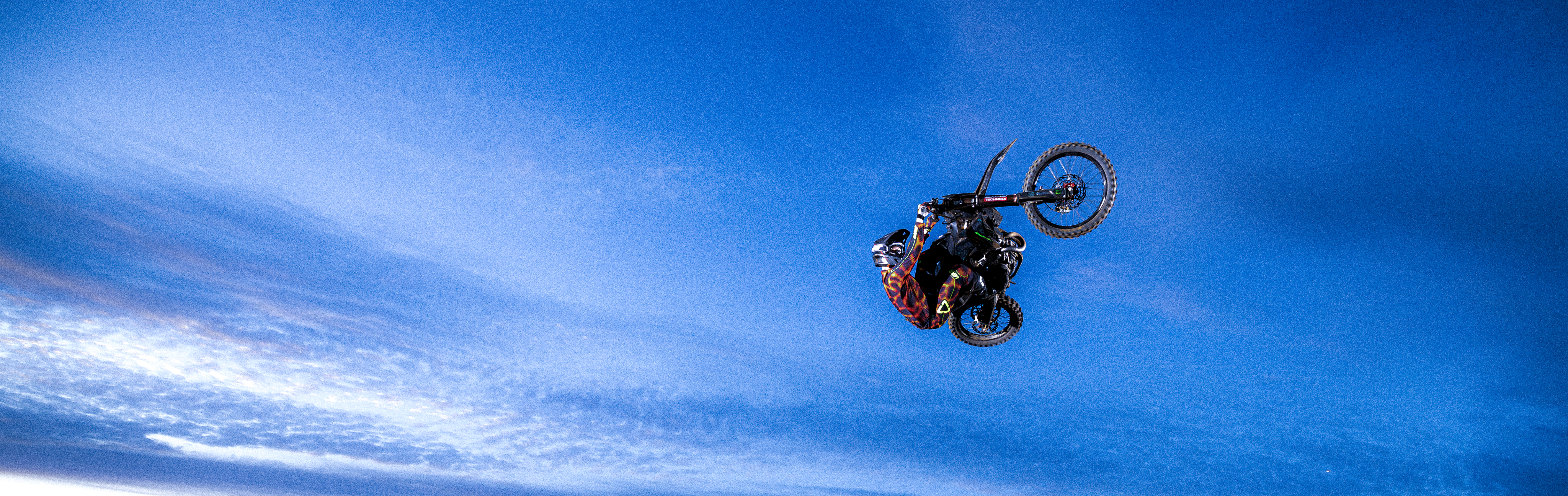 A motocross rider in a helmet and suit jumps a dirt bike mid-air against a bright blue sky with wispy white clouds.