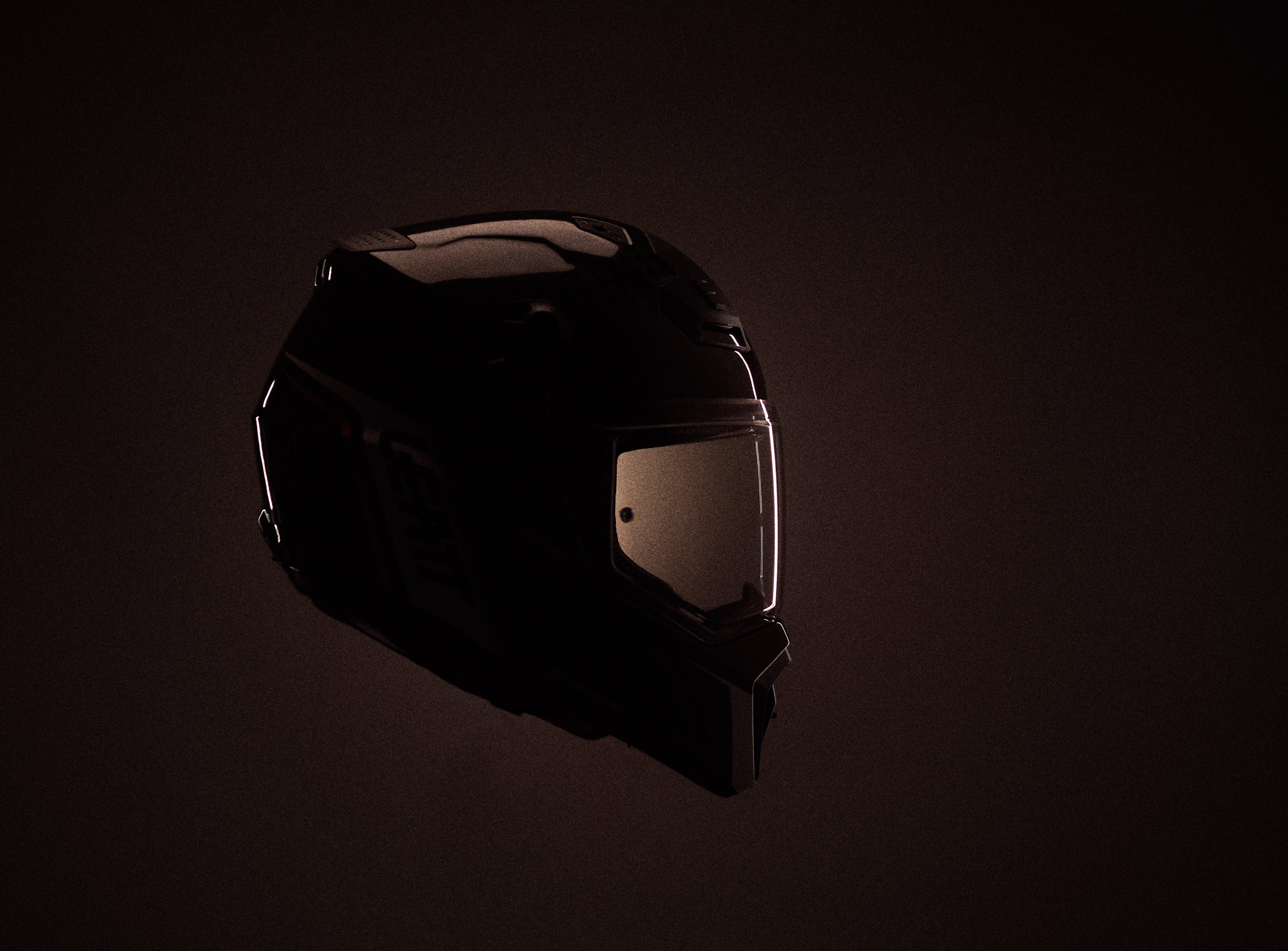 A dark full-face motorcycle helmet shown in dramatic low light against a very dark brown background, with the visor area illuminated slightly from the side.