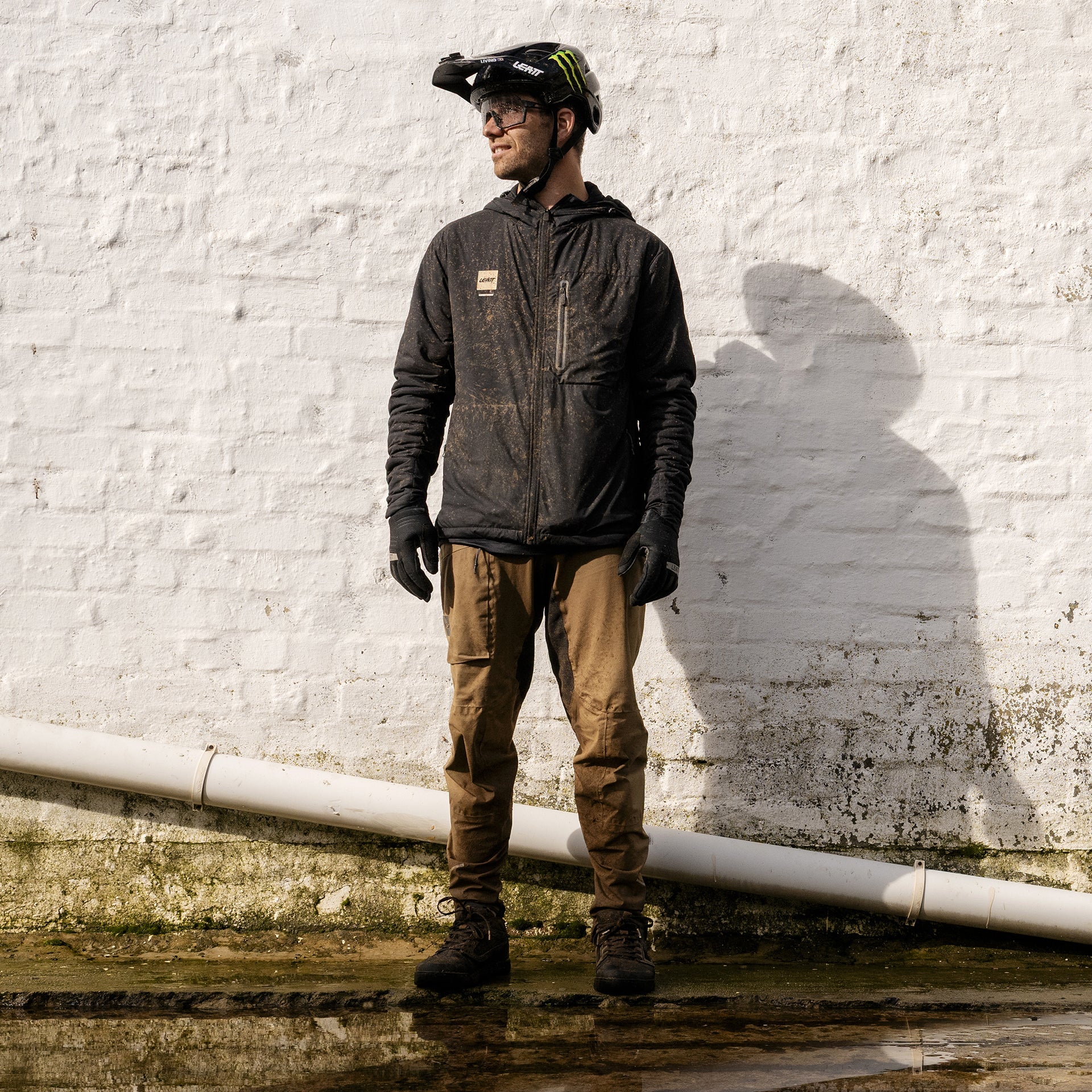 Person wearing a dark jacket and brown pants standing against a white brick wall.