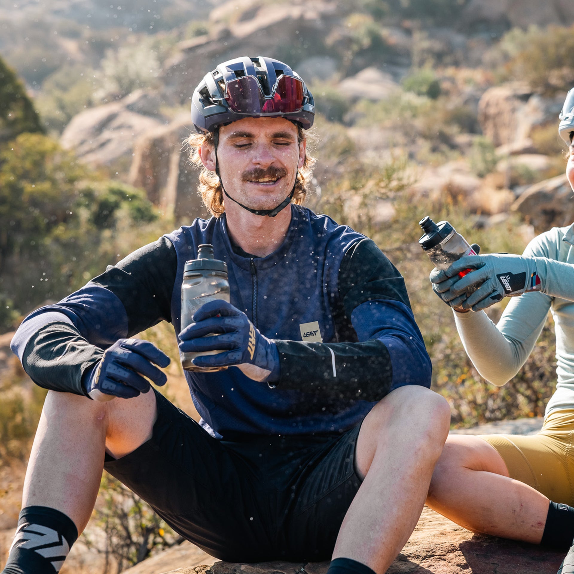 Two cyclists taking a break on a rocky trail, holding water bottles.