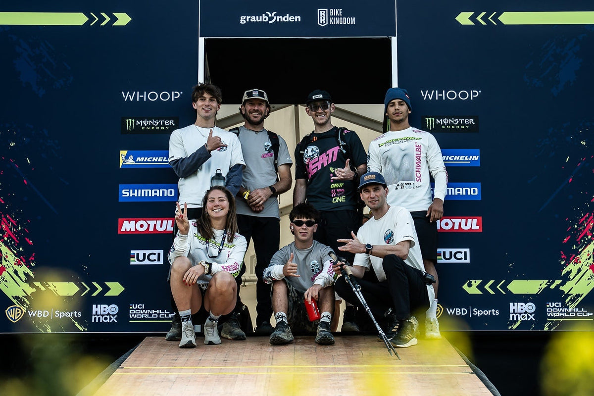 Six athletes posing together on a wooden ramp in front of a large dark blue Whoop Lenzerehide event backdrop with sponsor logos.
