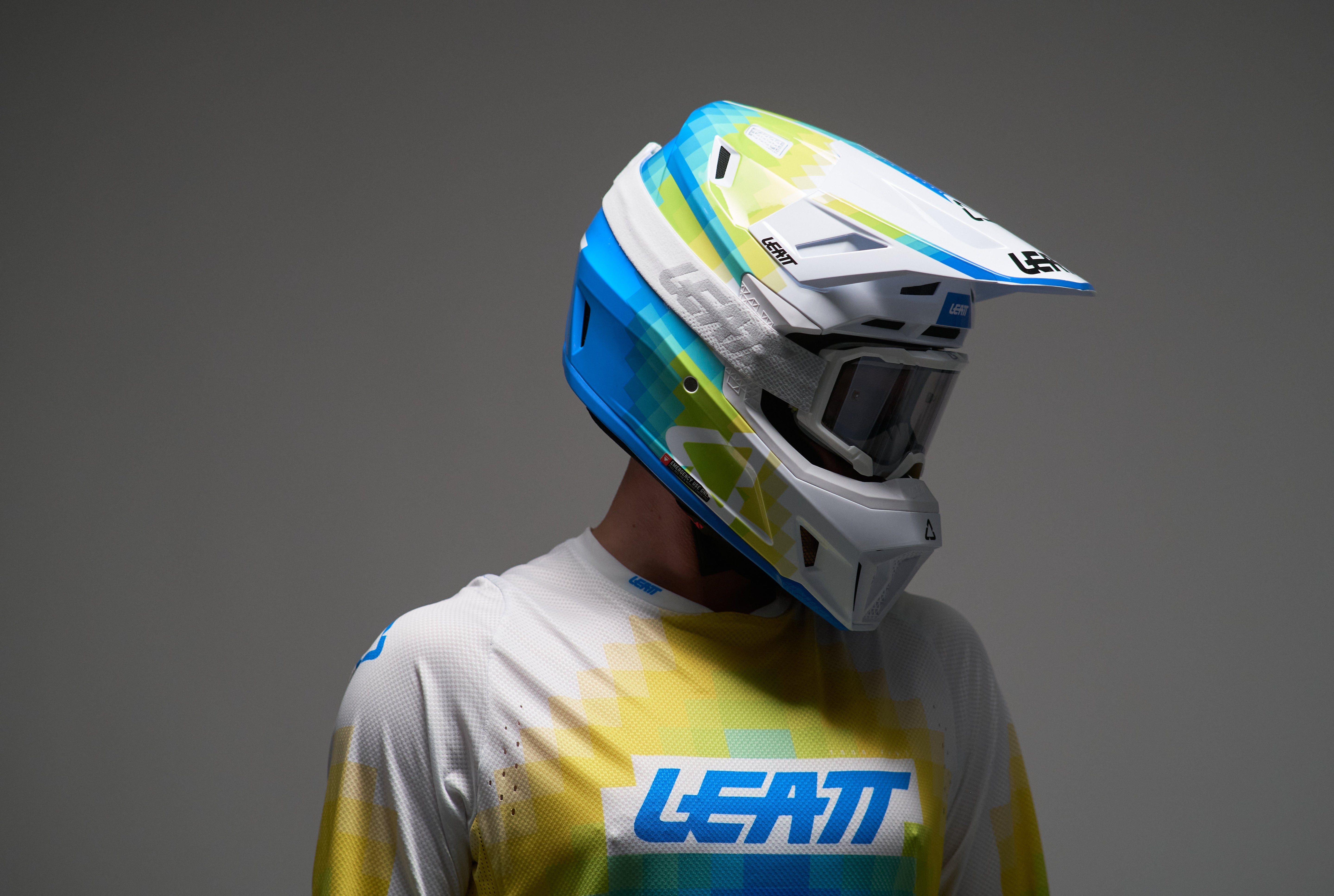 Person wearing a colorful motocross helmet and Leatt jersey on a gray background